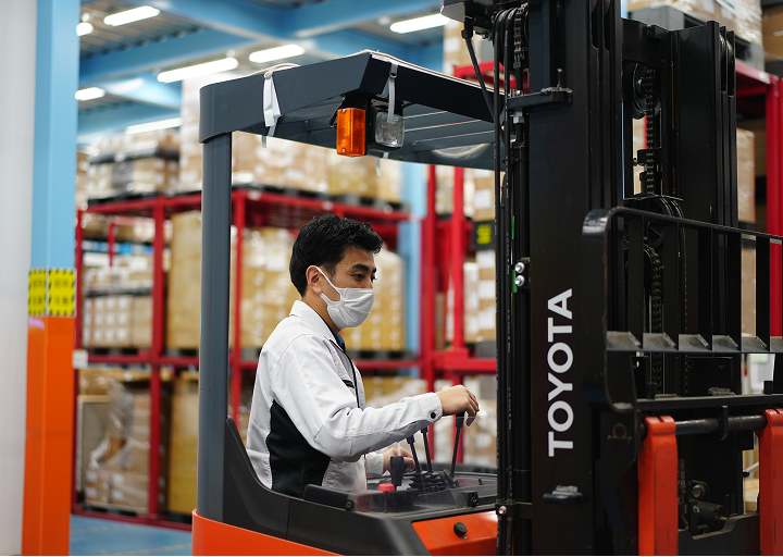 倉庫内でマスクを着けた作業員がフォークリフトを操作し、棚の荷物を運搬している様子