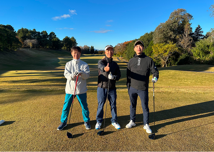 青空の下、ゴルフ場のフェアウェイで3人の男性がクラブを手に並んで立つ姿